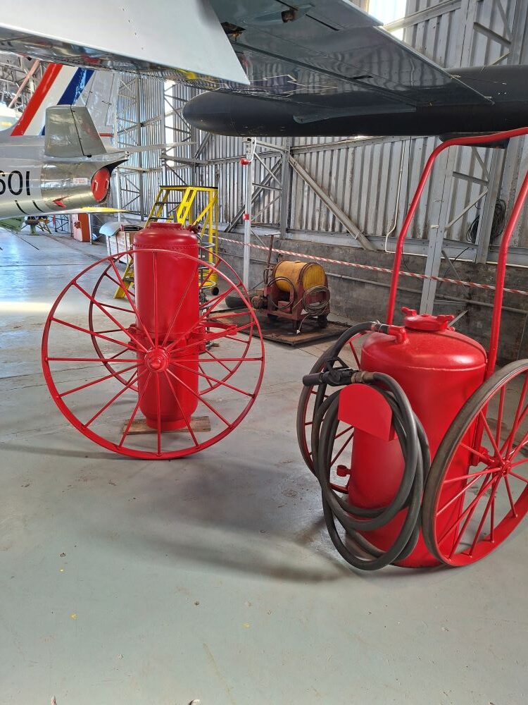 Two vintage red mobile airfield fire extinguishers on large spoked wheels displayed inside the hangar at the South African Air Force Museum during a Wild Buggers classic car club run.