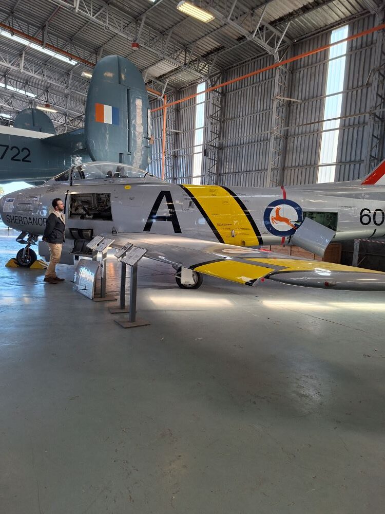 A vintage silver Canadair Sabre jet fighter named Sherdanor with yellow stripes on display inside the hangar at the South African Air Force Museum during a Wild Buggers club run for custom beach buggies, Baja Bugs, and classic VW air-cooled vehicles.