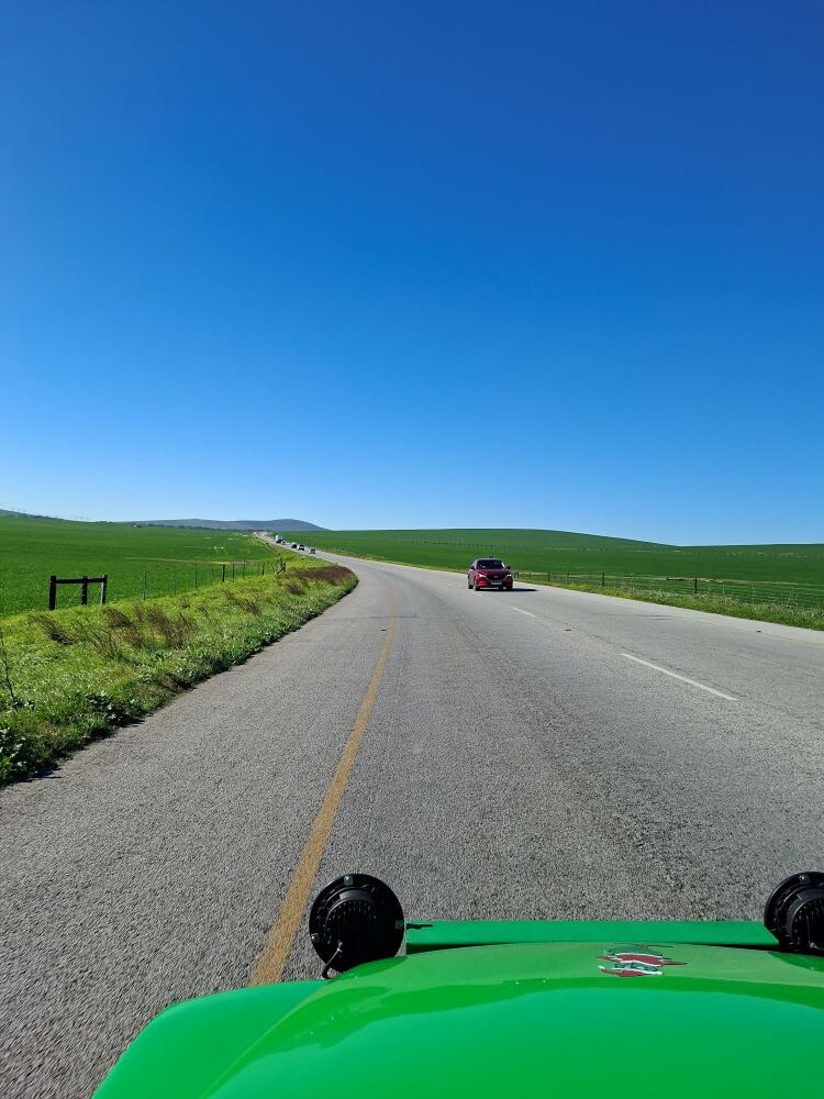A view from inside a bright green custom beach buggy driving along a scenic open road through lush green fields under a clear blue sky during a classic car club run.