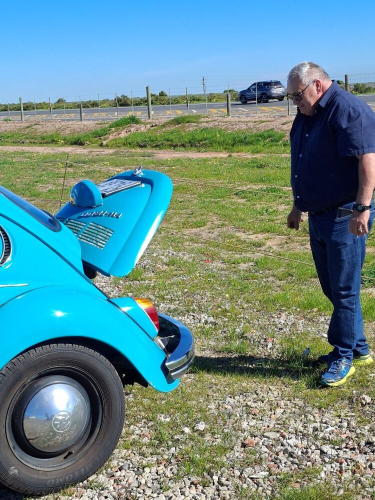 A close-up view of a club member looking at the open engine lid of a turquoise blue classic VW air-cooled Beetle during a maintenance stop on a Wild Buggers club run featuring custom beach buggies and Baja Bugs.