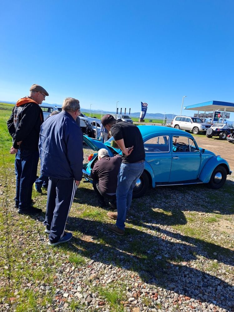 A group of club members gathered around to troubleshoot the overheating engine of a turquoise blue classic VW air-cooled Beetle during a stop on the Wild Buggers Propellers and Pistons Winter Cruise.