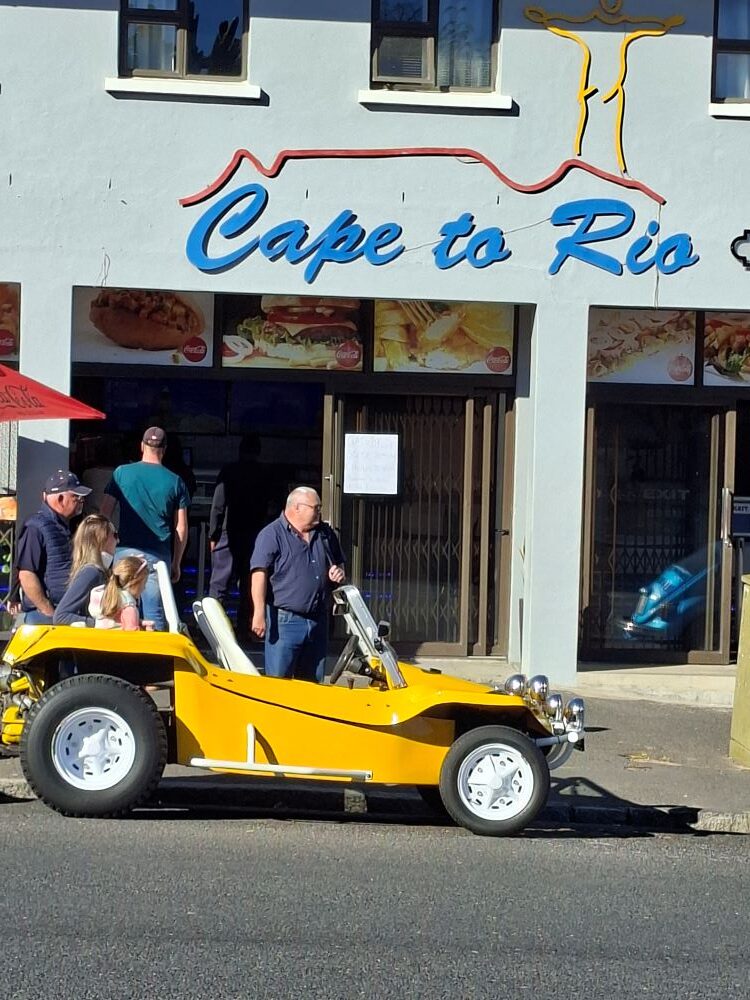 Club members Herbie and Boeta standing next to a bright yellow custom beach buggy parked in front of Cape to Rio Fisheries during the Propellers and Pistons Winter Cruise, an event celebrating classic VW air-cooled vehicles and Baja Bugs.