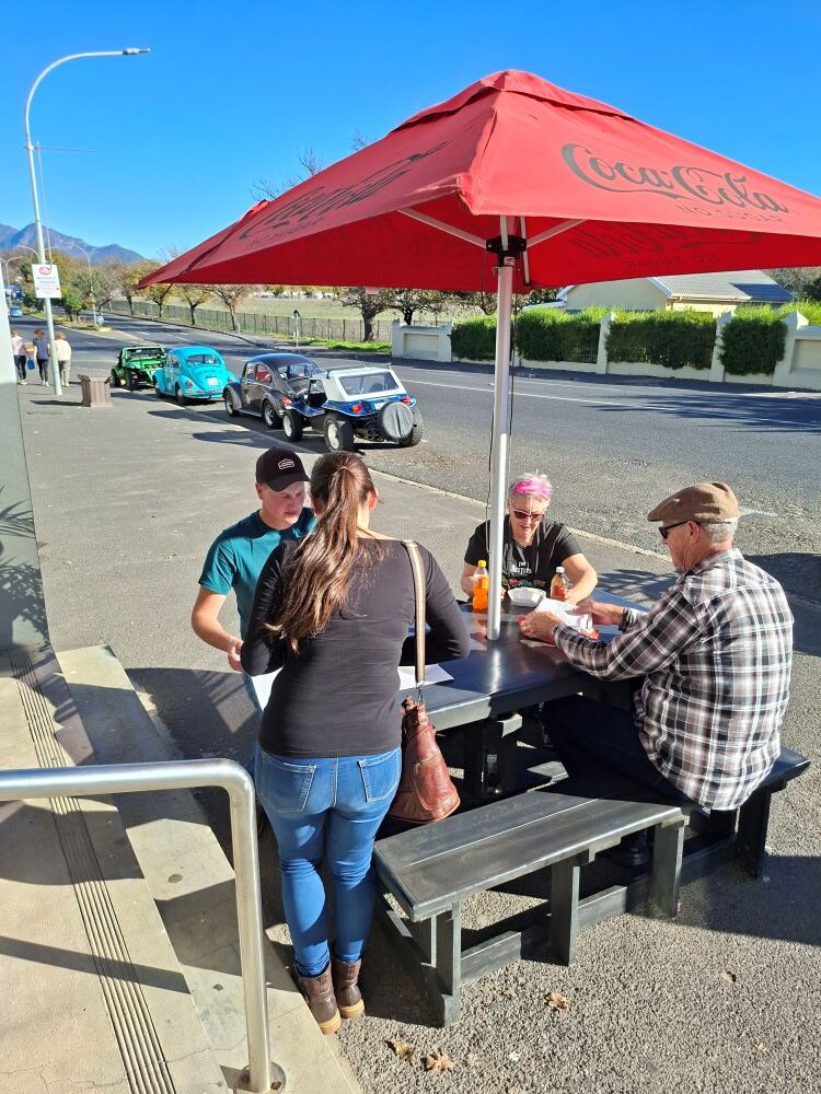 Club members enjoying an outdoor meal at Cape to Rio Fisheries during the Propellers and Pistons Winter Cruise, with a convoy of custom beach buggies, Baja Bugs, and classic VW air-cooled vehicles parked in the background.
