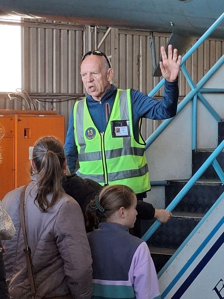 A museum tour guide wearing a high visibility vest addressing visitors inside a hangar at the South African Air Force Museum during a car club run.