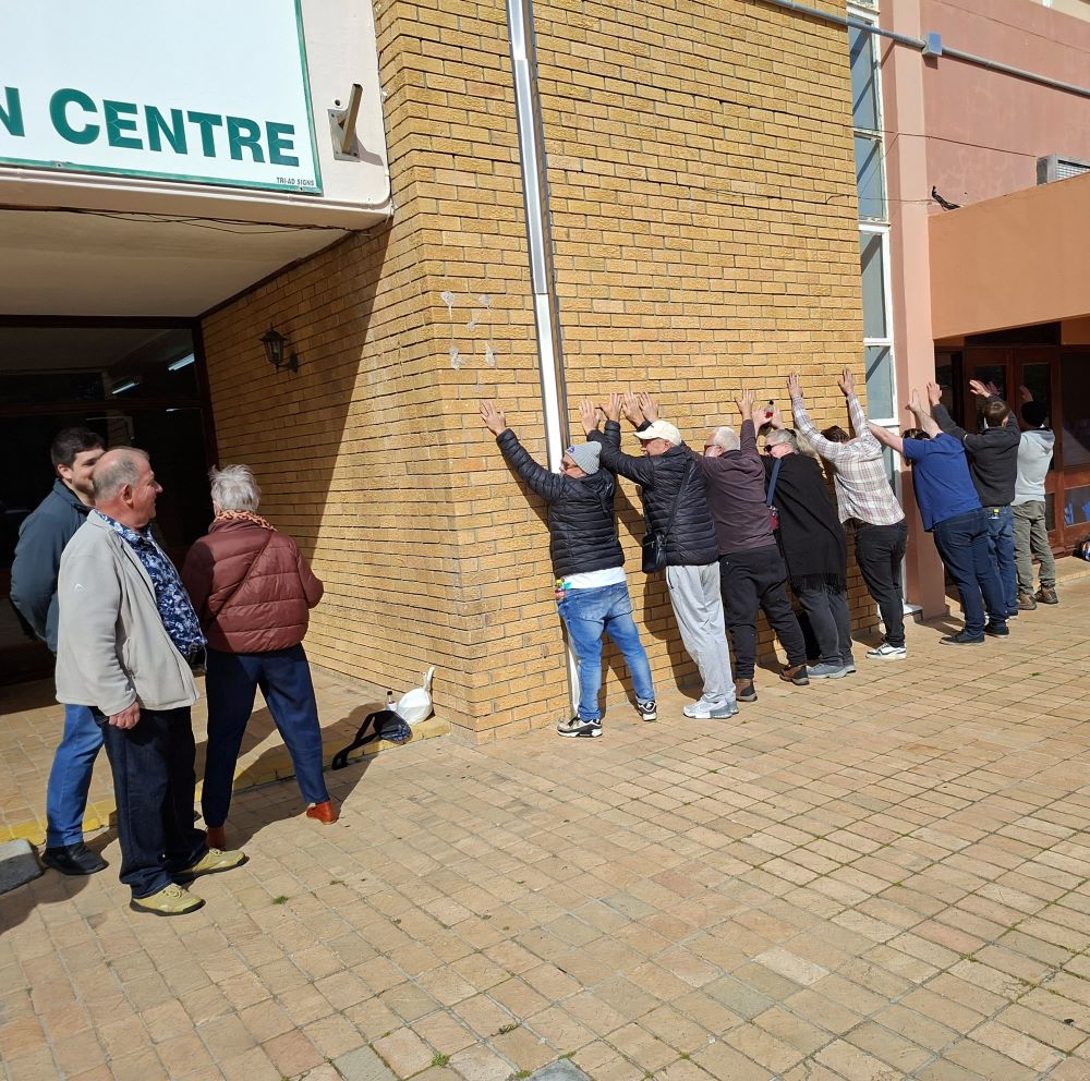 Wild Buggers club members posing with hands against the brick wall outside the Pollsmoor brunch stop during the From Bars to Battleships run, surrounded by classic VW air-cooled enthusiasts.