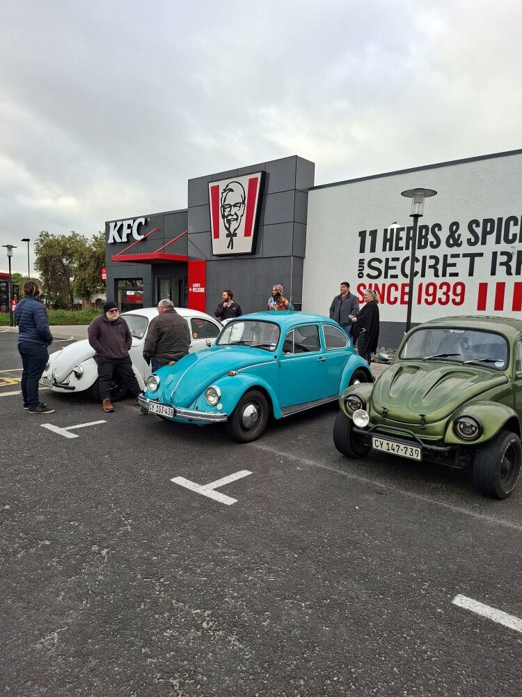 Club members chatting beside a classic VW Beetle, Baja Bug, and air-cooled vehicles at the morning meetup in Bothasig before the From Bars to Battleships run.