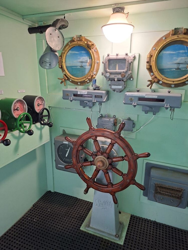 A ship's wheel, telegraph dials, and brass portholes inside a simulated ship bridge exhibit at the museum.