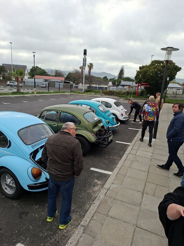 Club members inspecting engines and discussing custom modifications on a Beach Buggy, Baja Bug, and classic VW air-cooled vehicles at the meetup spot before the From Bars to Battleships run.