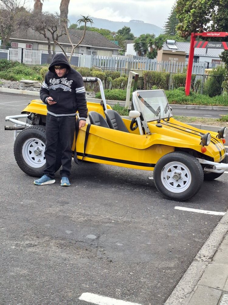 Wiehann Geldenhuys standing next to his yellow Beamish Beach Buggy, 'Bugg', looking at his phone while organizing the upcoming Wild Buggers club run in the Western Cape.