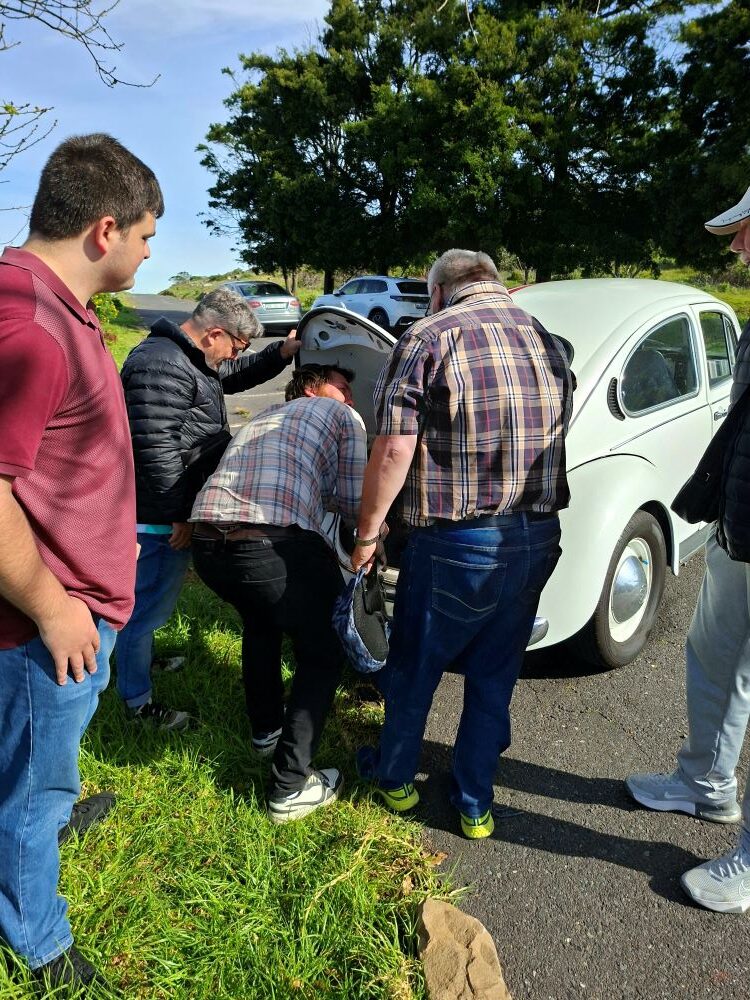 Club members gathered around the engine bay of a classic VW Beetle to troubleshoot a roadside mechanical issue.