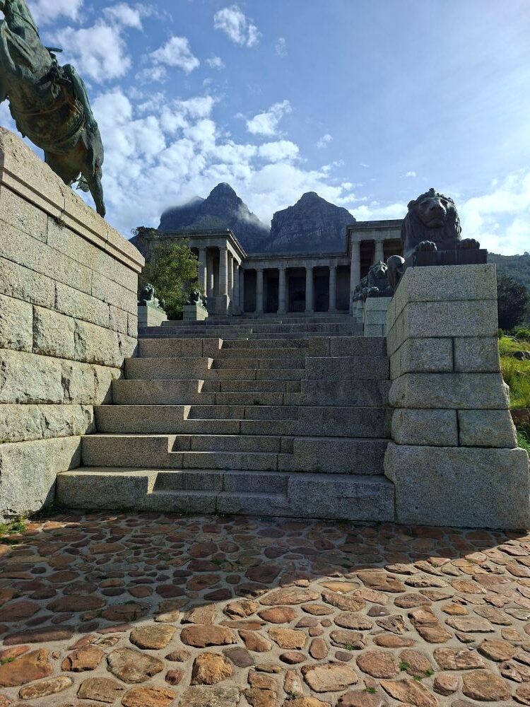 The stone steps and columns of Rhodes Memorial leading up under a bright blue sky with mountain peaks in the background.
