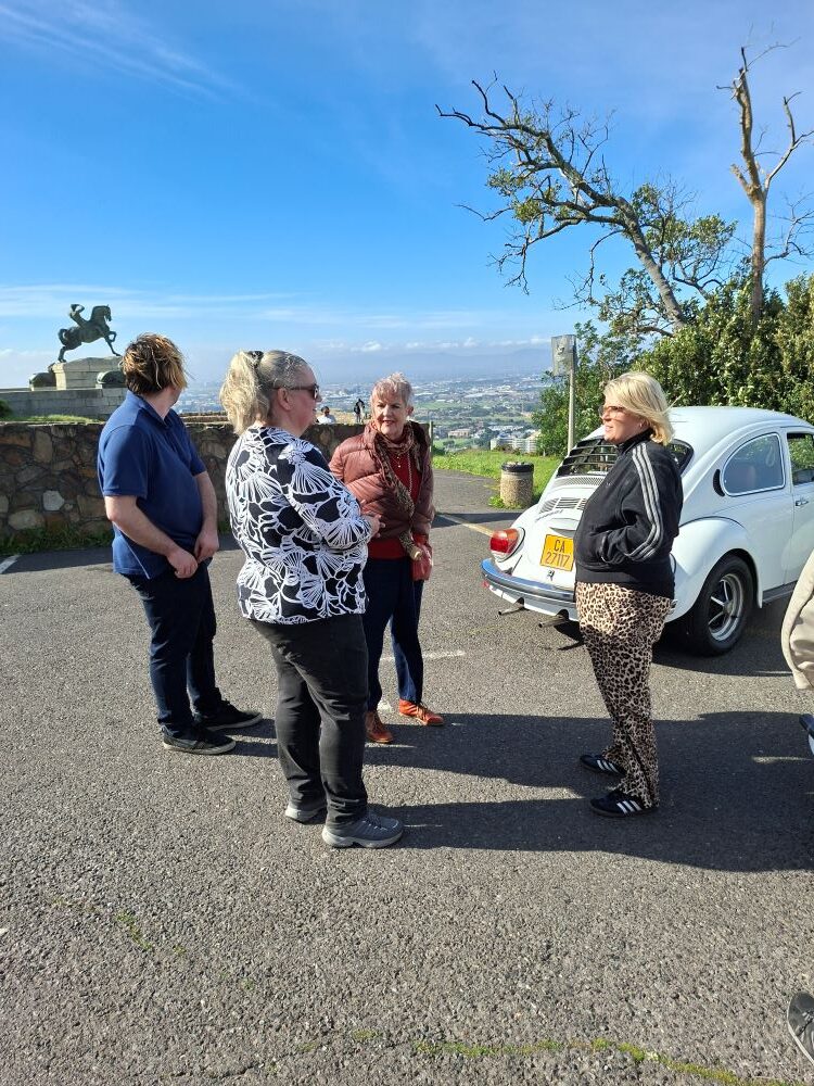 Club members chatting and relaxing near a white classic VW Beetle at Rhodes Memorial with scenic views in the background.