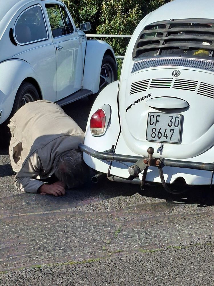 A person inspecting the underside of Bertus's white classic VW Beetle parked on the roadside during the club run.