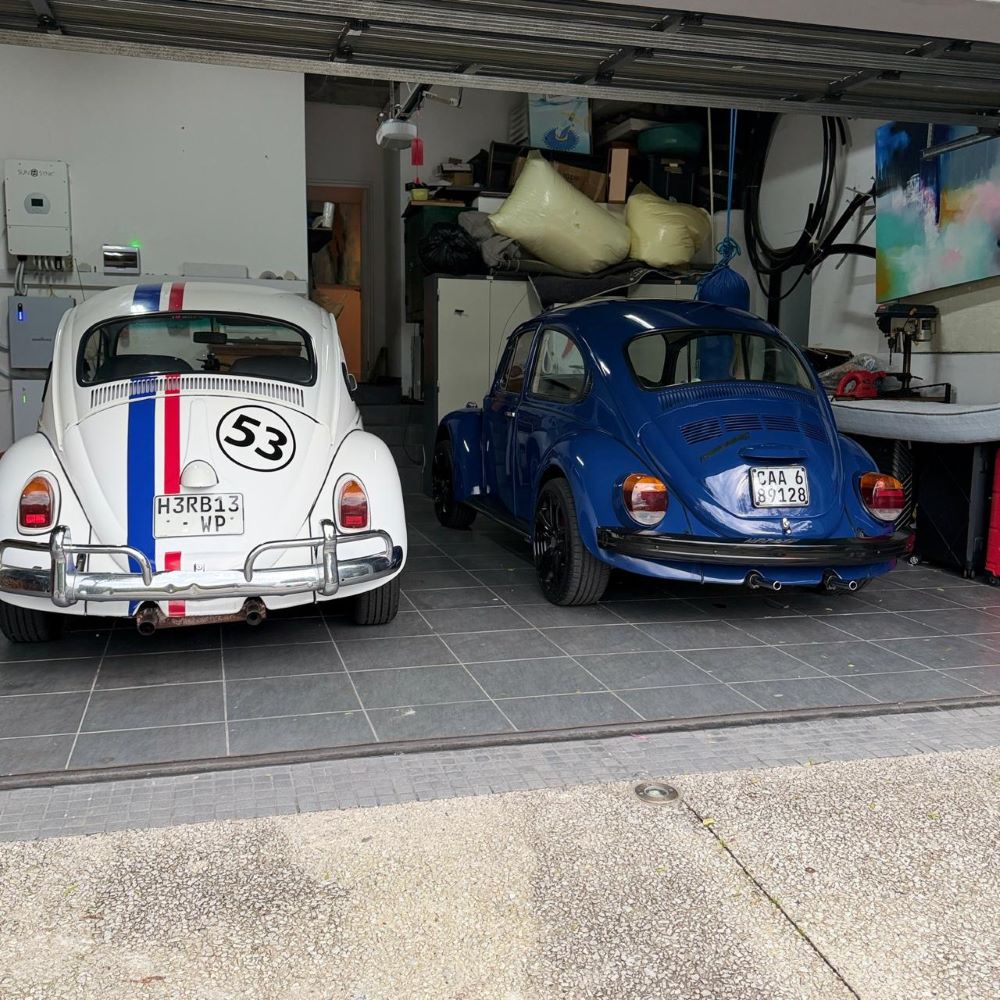 Neil's Herbie classic air-cooled VW and Hans's newly restored blue VW Beetle called Thunder Bug parked side by side in a garage.