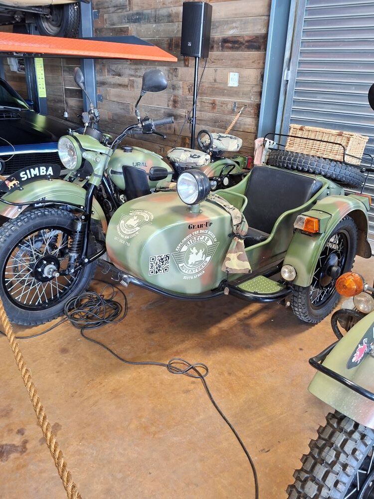 A rugged green Ural sidecar motorcycle parked indoors alongside other vintage vehicles during the club stop.