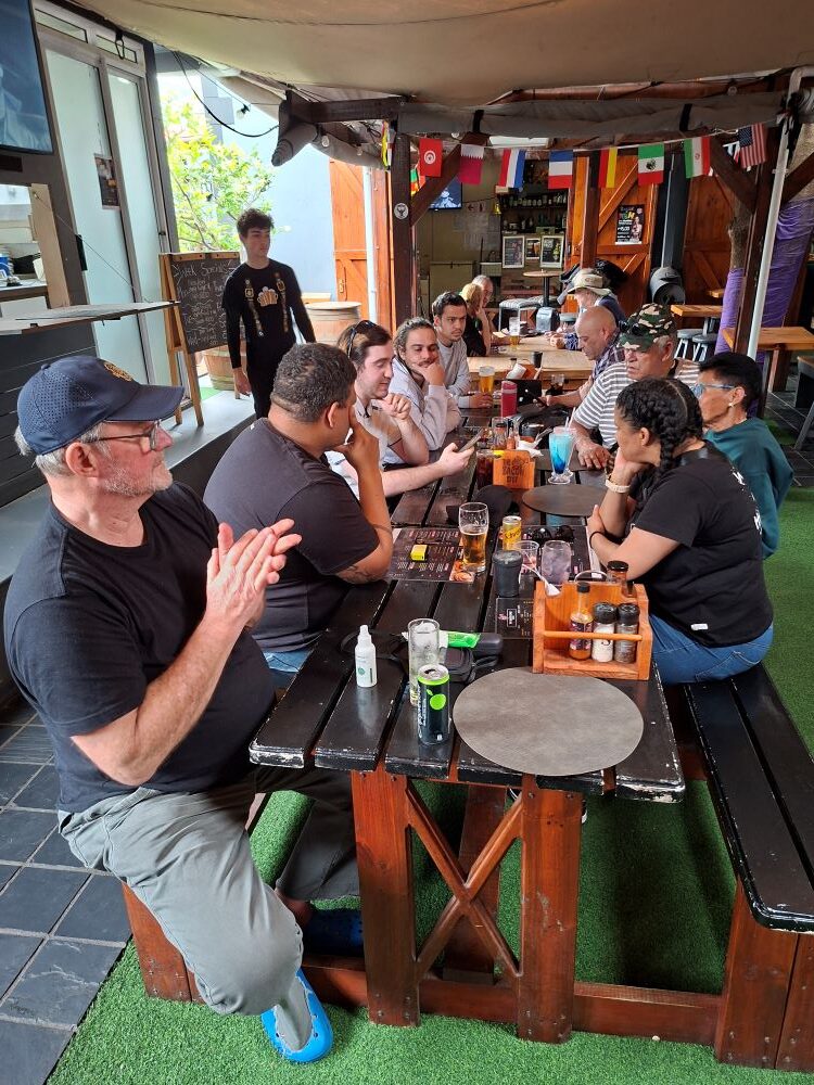 A wide shot of club members relaxing around large wooden tables at the restaurant during the convoy stop.