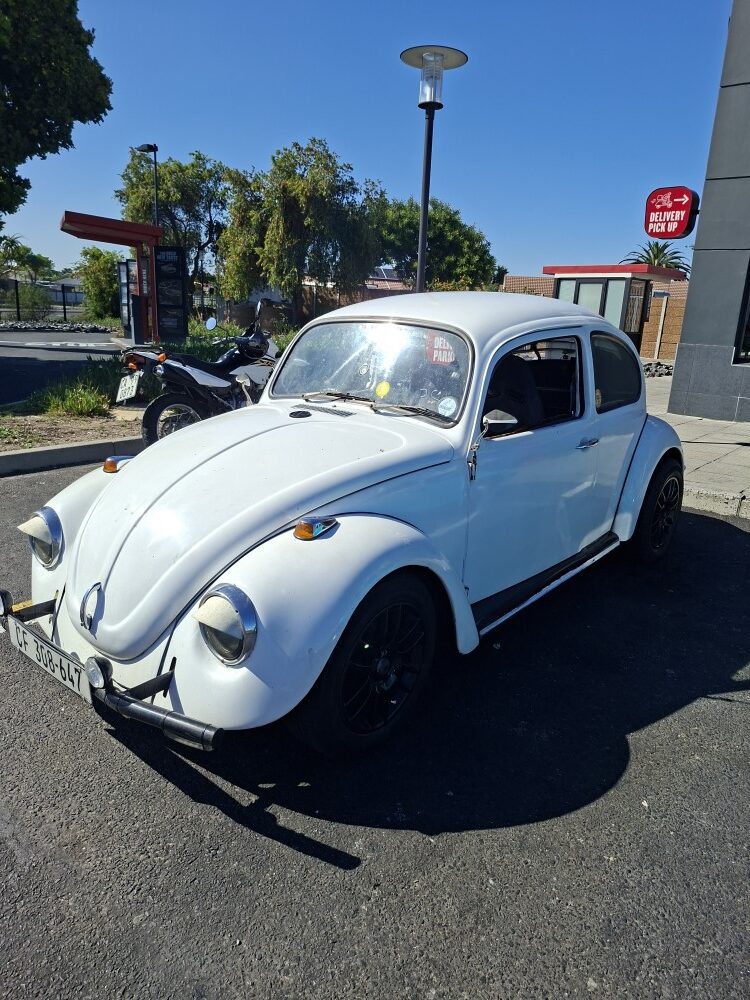 A white classic VW Beetle with black alloy wheels and headlight eyelids parked at the Bothasig KFC meetup.