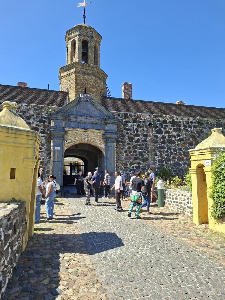A group of people walking through the stone archway entrance of the historic Castle of Good Hope under a clear blue sky.