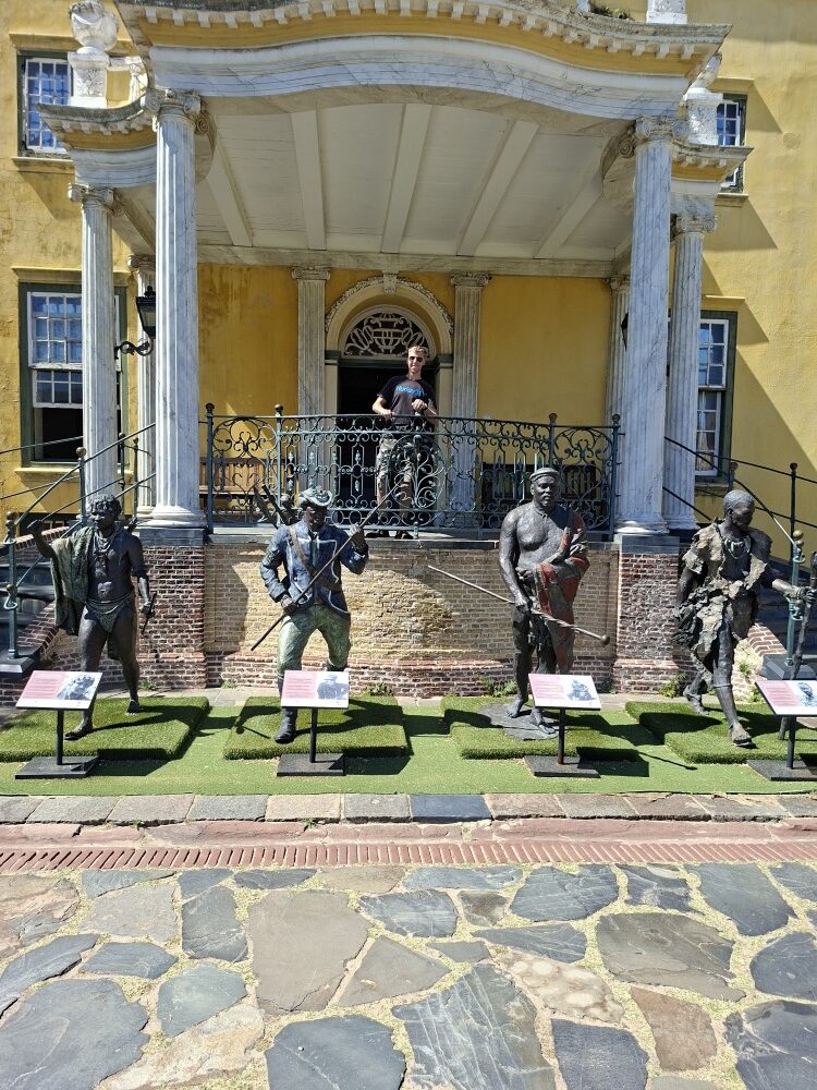 A man standing on an elevated portico behind four life-size historical bronze statues in a cobbled courtyard at the Castle of Good Hope.