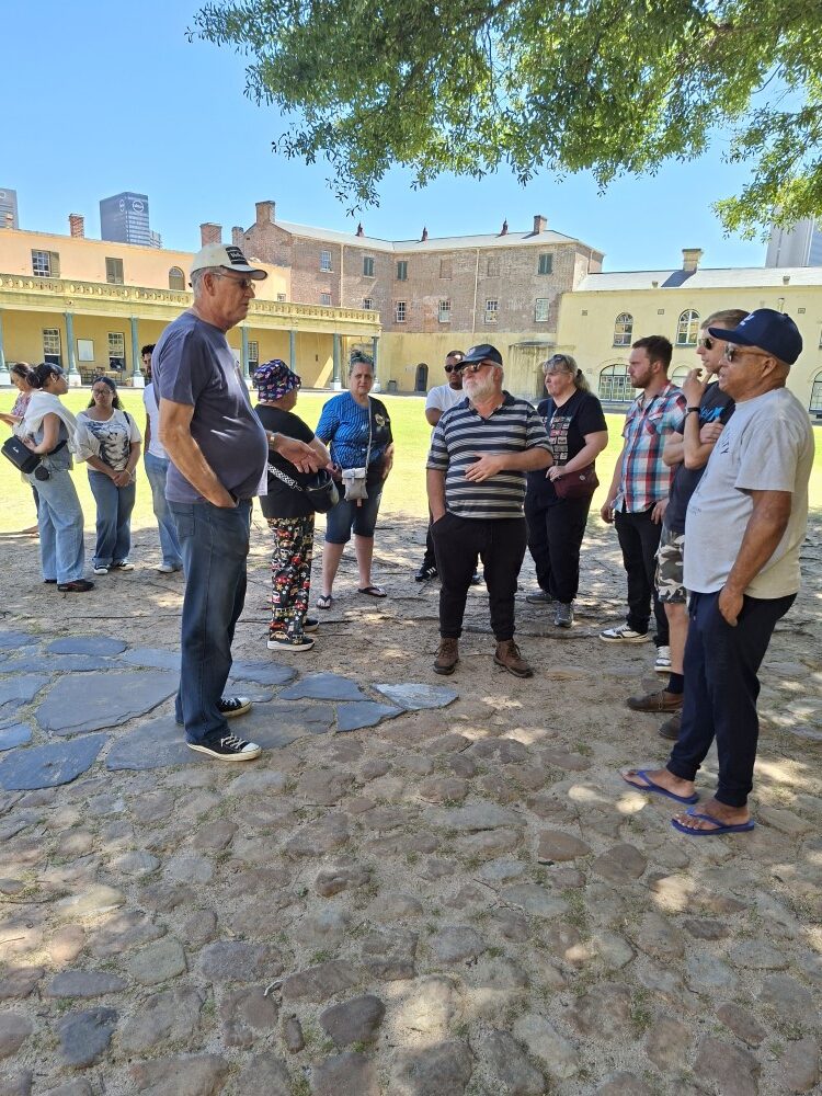 A group of club members standing in a circle in the cobblestone courtyard of the Castle of Good Hope under tree shade.