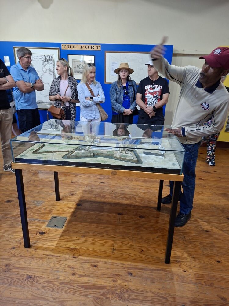 A tour guide demonstrating details over a glass display model of the historic fort to a group of attentive visitors.