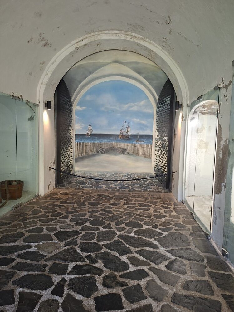 A stone-paved vaulted archway with large iron gates featuring a realistic mural of historical tall ships sailing in Table Bay.