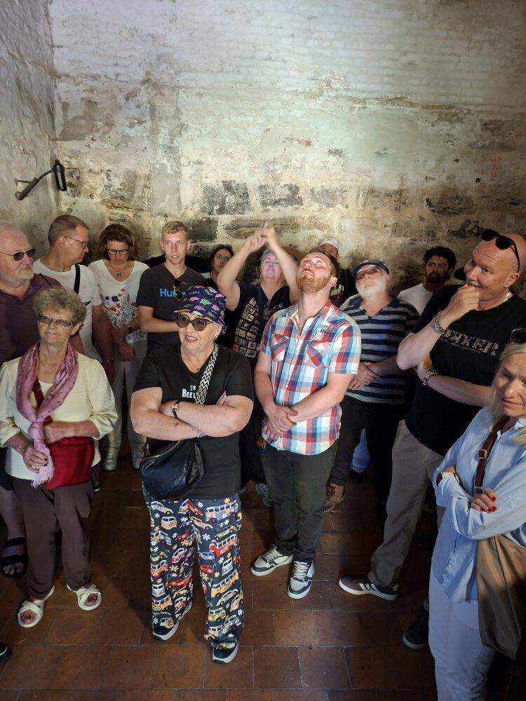A group of people standing close together within a dimly lit, rough stone-walled dungeon room during a guided historical tour.