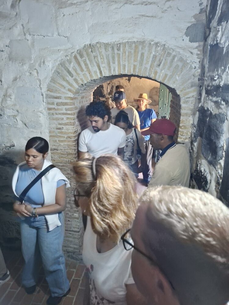 A group of visitors walking single file through a low brick arched doorway into a stone dungeon room.
