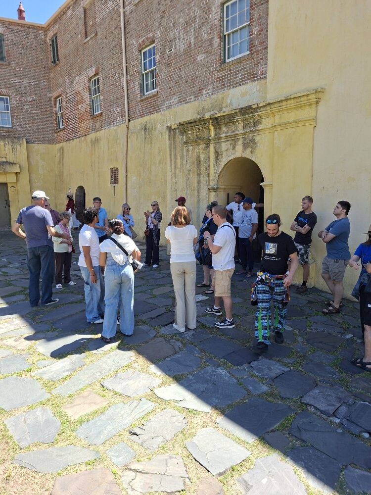 A tour group gathered on paved stone flags outside a historic yellow brick building during a guided walkthrough.