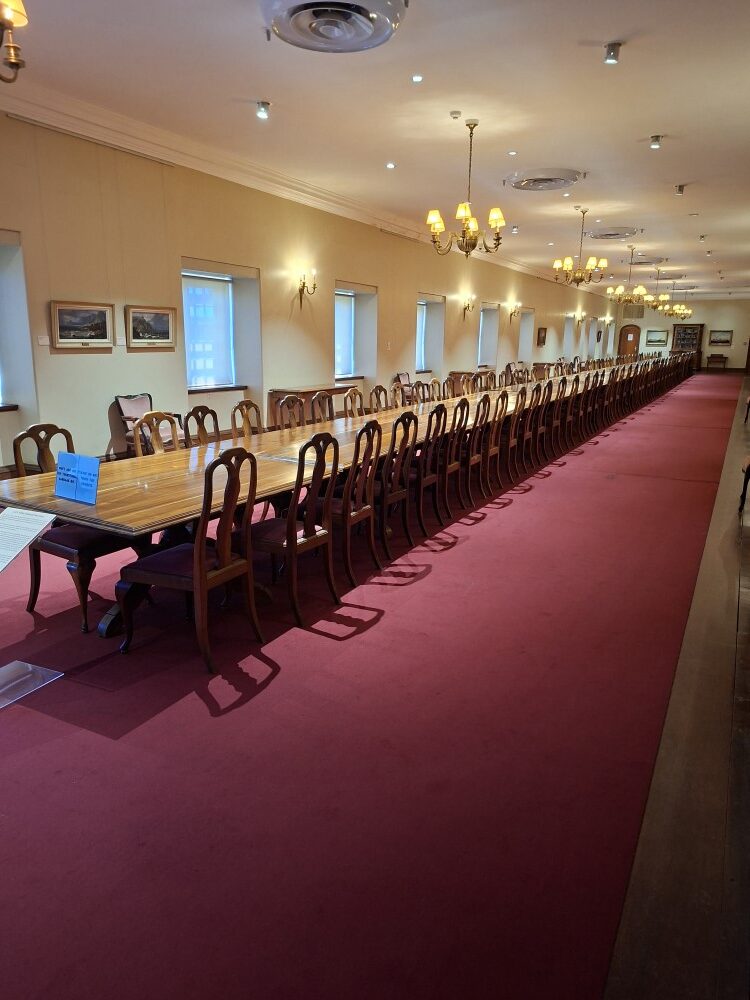 An exceptionally long polished wooden dining table with dozens of matching chairs lining a carpeted hall with chandeliers inside the Castle.
