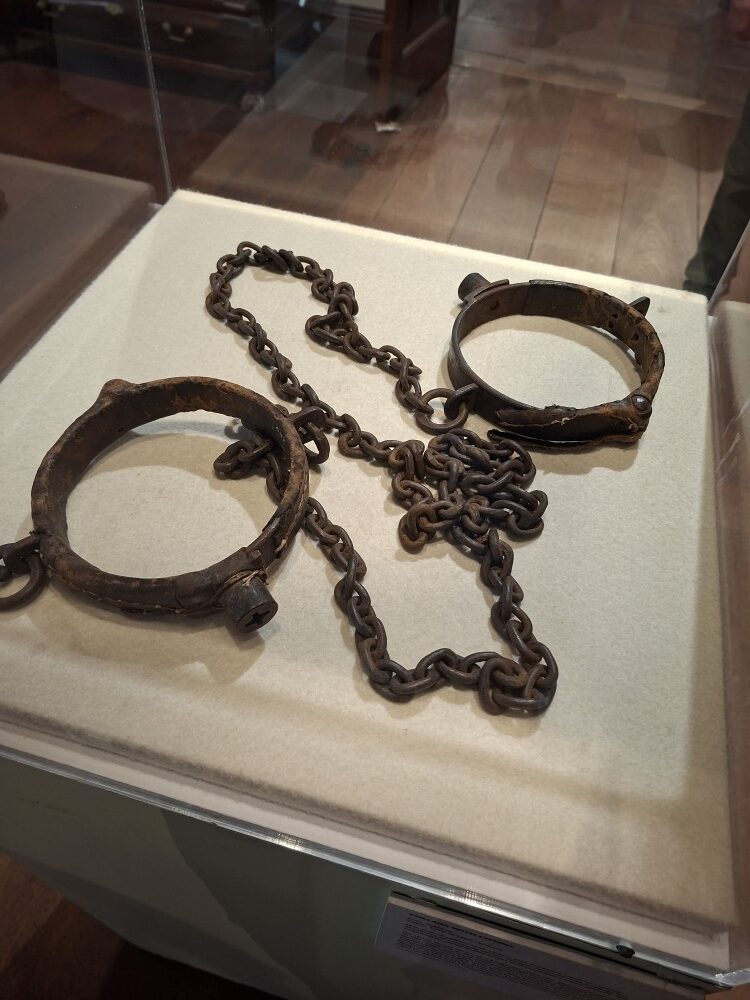 Heavy weathered iron prisoner leg shackles and connecting chains displayed inside a glass museum case.