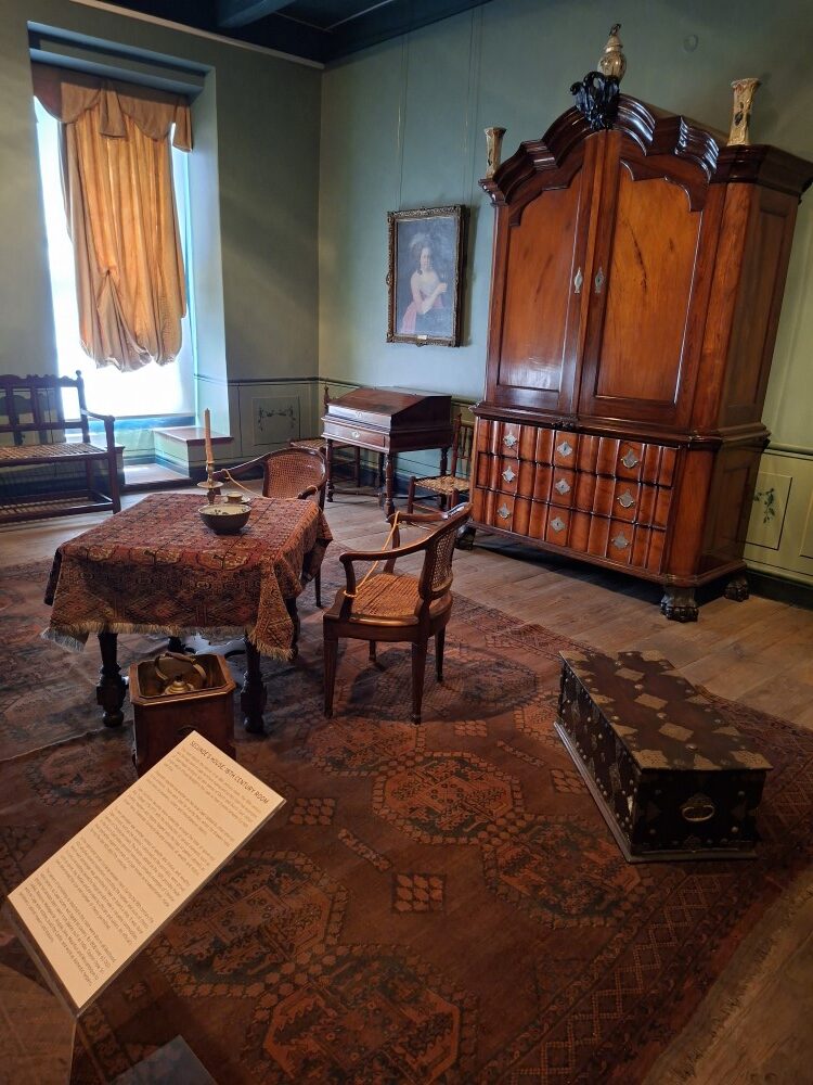 An 18th-century restored room exhibit featuring vintage wooden furniture, a large armoire, woven carpets, and an information display board inside the Castle museum.