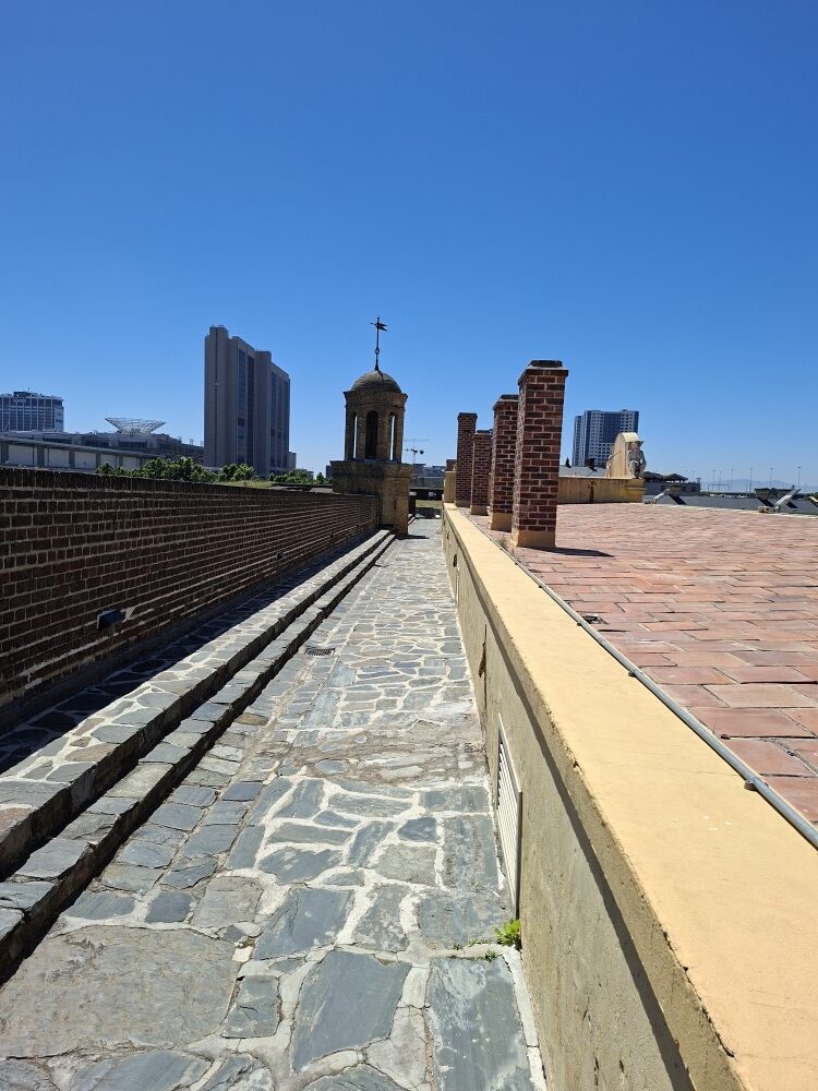 A high elevated stone walkway on top of a historic fort wall with brick pillars and a small bell tower overlooking urban buildings under a clear blue sky.