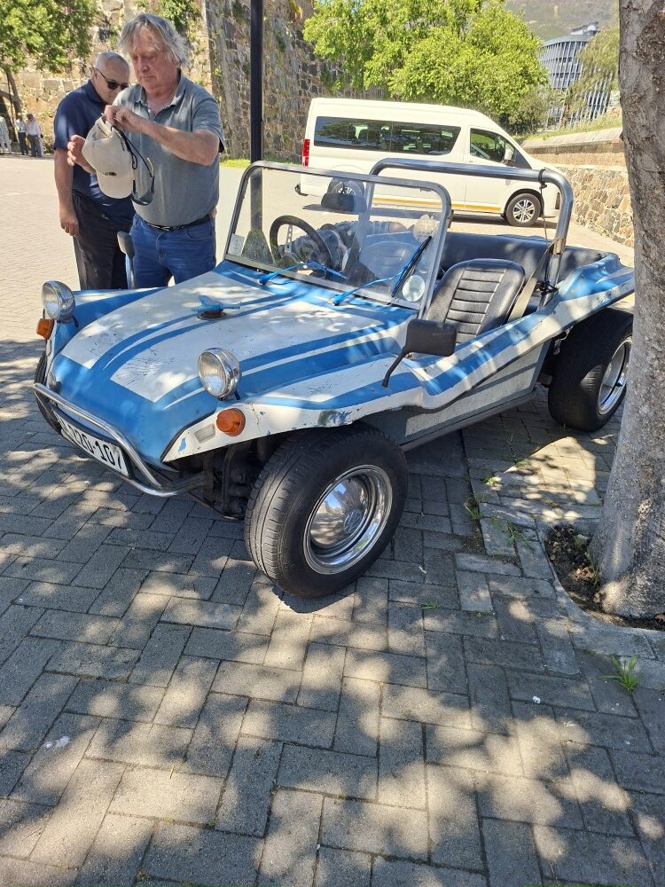 Harvey Metcalf standing beside his blue and white Beamish beach buggy in a paved parking lot under tree shade.