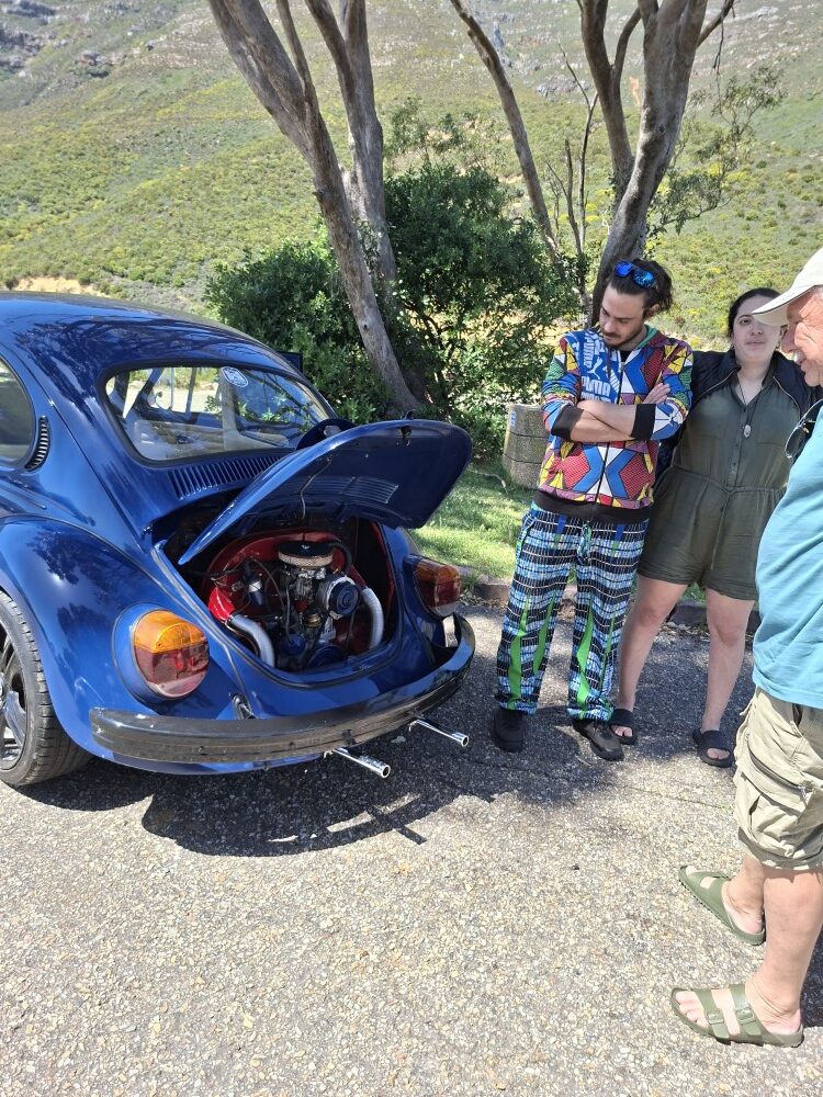 Club members standing around a dark blue vintage VW Beetle with an open rear engine lid at a roadside picnic area.