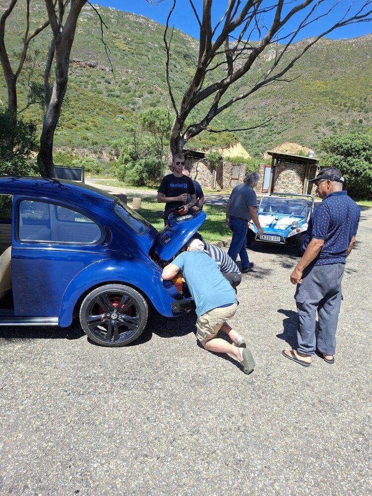 Club members leaning over the rear engine bay of a blue classic VW Beetle in a sunny gravel parking area.