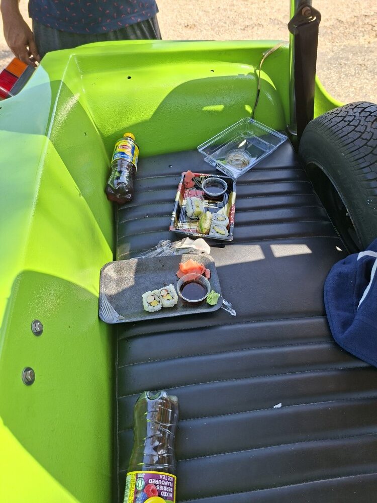 Sushi platters and iced tea bottles resting on the black vinyl back seat of a bright green beach buggy during a picnic stop.
