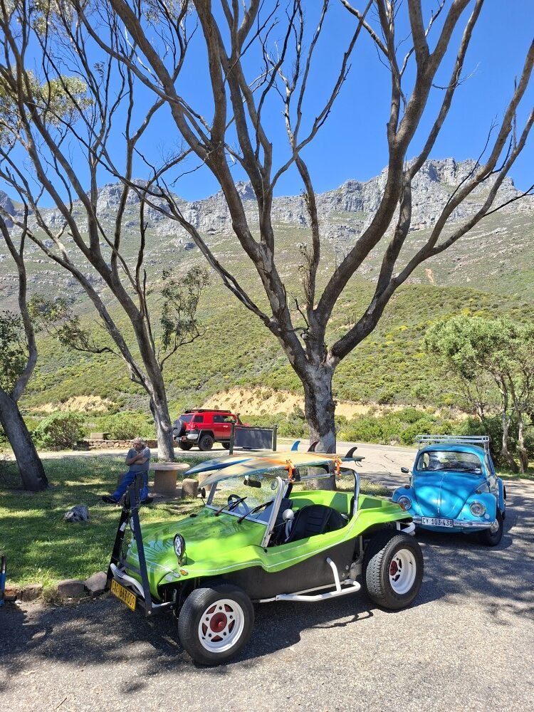 A bright green beach buggy with surfboards strapped to the roll cage parked beside a blue classic Beetle at a mountain picnic stop.