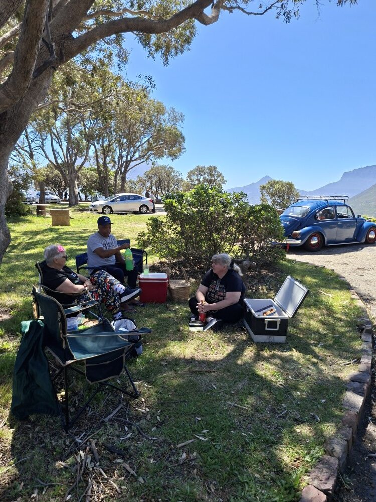 Club members sitting on camp chairs in the grass under trees enjoying a picnic next to a classic blue VW Beetle.