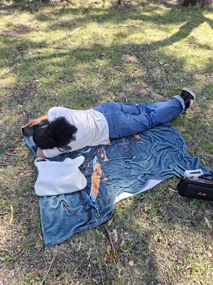 A person lying on a picnic blanket in the grass under tree shade during an outdoor club gathering.