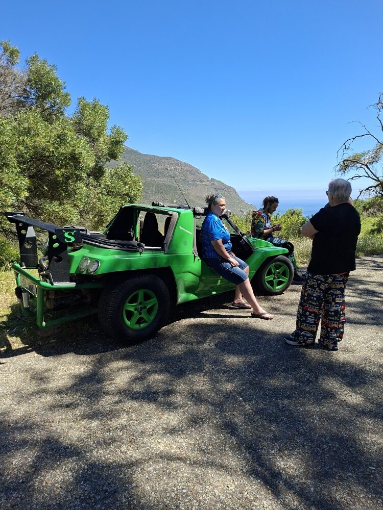 Mickey sitting on the side door frame of Shrek the green beach buggy chatting with club members overlooking coastal mountains.