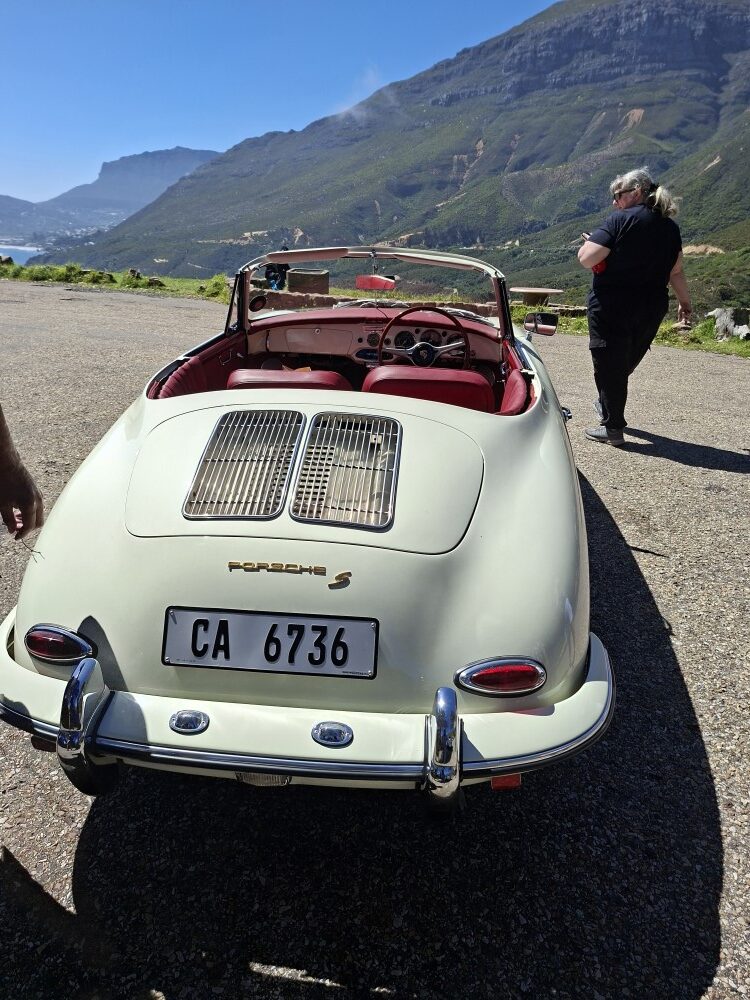 Rear view of a classic ivory Porsche 356 speedster convertible with red leather interior parked at a mountain lookout.