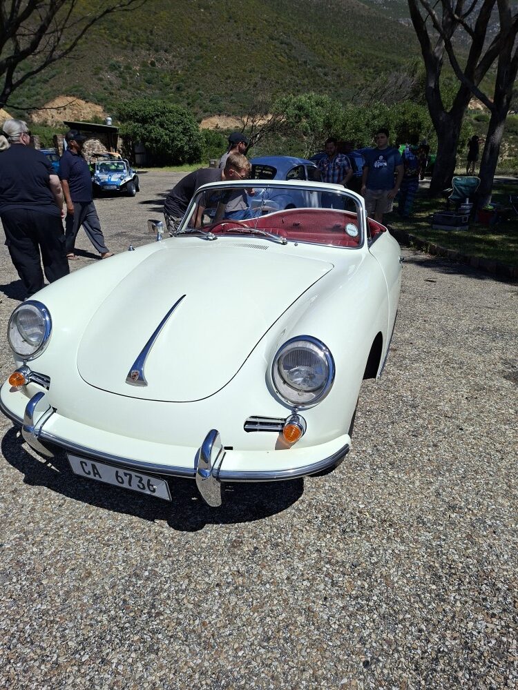 Front view of an ivory classic Porsche 356 convertible with chrome bumpers parked at a sunny gravel rest area.