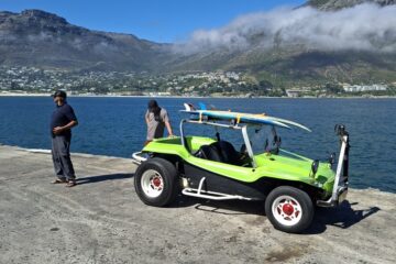 A bright lime green beach buggy with surfboards on the roll cage parked on the concrete wharf at Mariner's Wharf in Hout Bay overlooking the ocean and mountains.