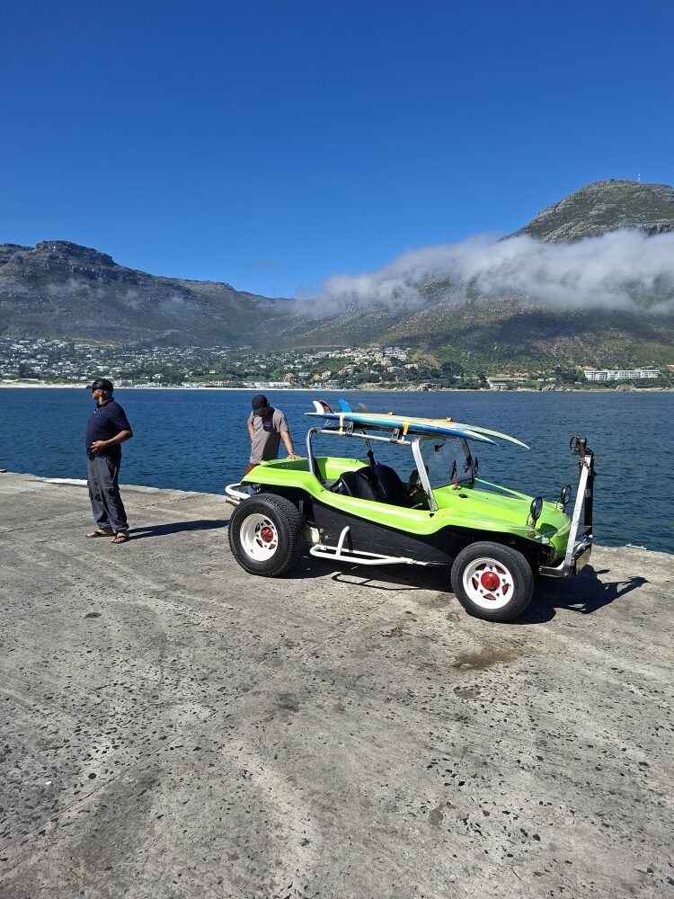 A bright lime green beach buggy with surfboards on the roll cage parked on the concrete wharf at Mariner's Wharf in Hout Bay overlooking the ocean and mountains.
