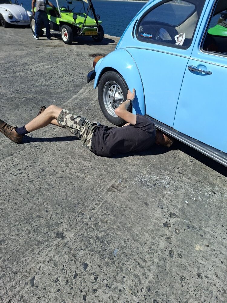 Kyle Cato lying on the harbour ground inspecting the underside and EMPI deep sump of his light blue classic VW Beetle.