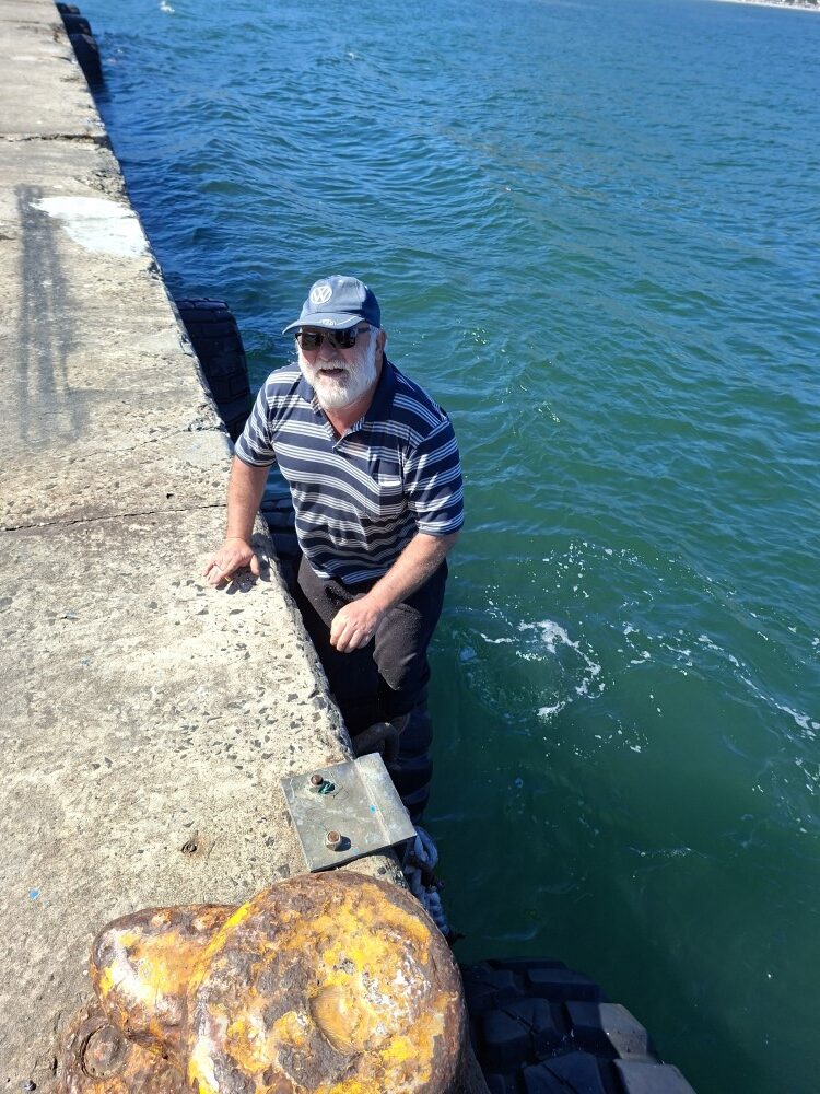 Leon Visser wearing a VW cap climbing inside a large harbour fender tire mounted to the edge of a concrete pier over deep blue ocean water.