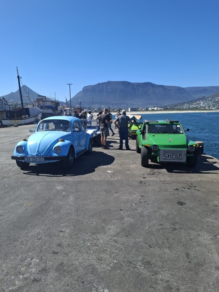 A light blue classic VW Beetle and the green beach buggy Shrek parked side by side on a concrete pier with members standing and chatting nearby.