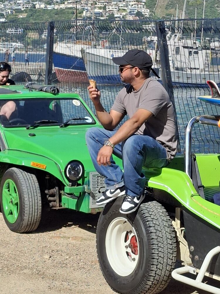 A club member wearing a cap and sunglasses sitting on the large rear tire of a green beach buggy enjoying an ice cream cone at the harbour.