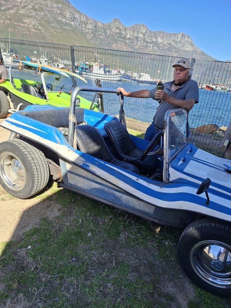 Harvey Metcalf wearing a cap leaning against the roll cage of his blue and white striped beach buggy at the harbour parking lot.